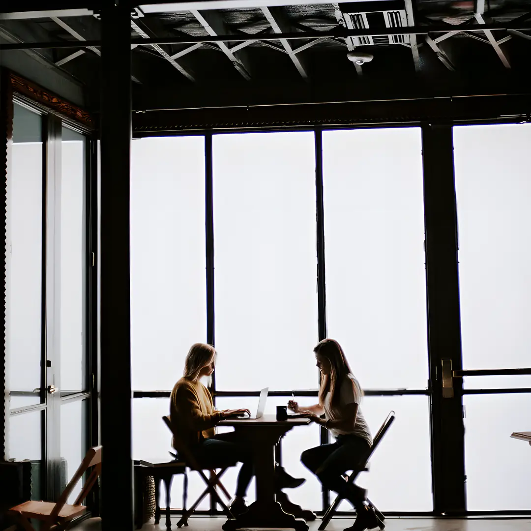 Two colleagues working at a table beside tall windows in a loft studio