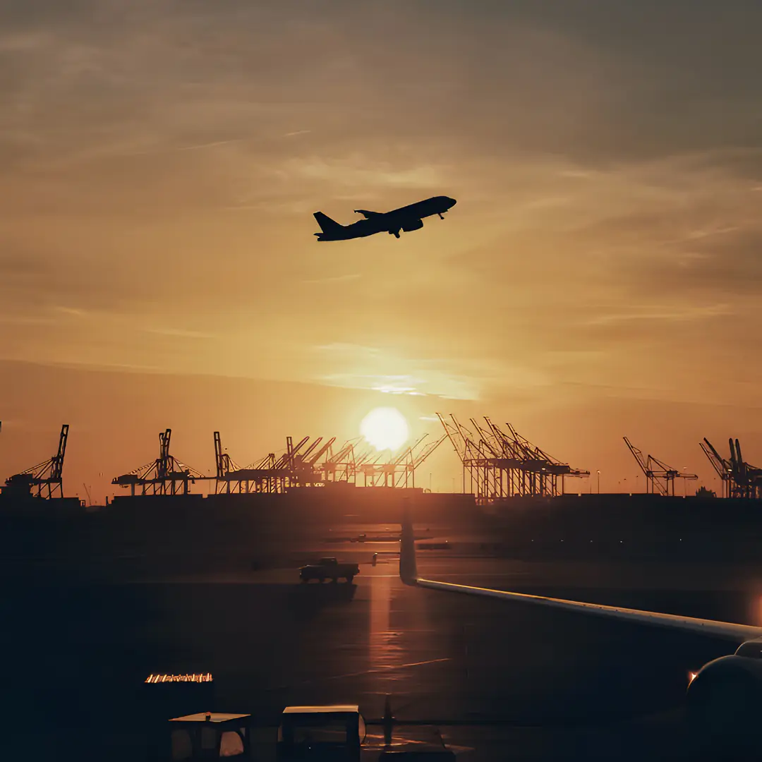 A plane taking off at sunset above port cranes