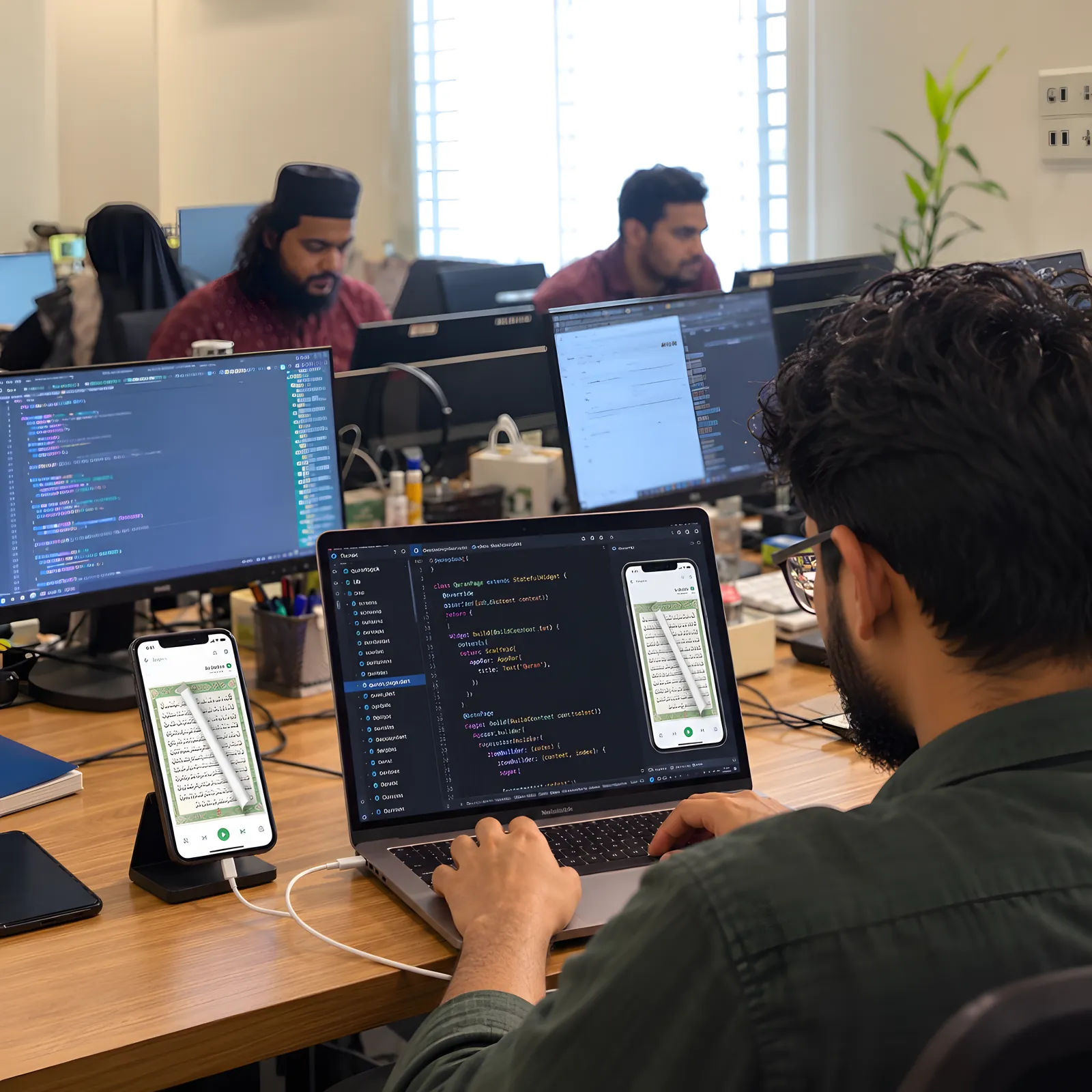A developer coding the Quran Touch app on a laptop with the app running on a phone beside him in the team office