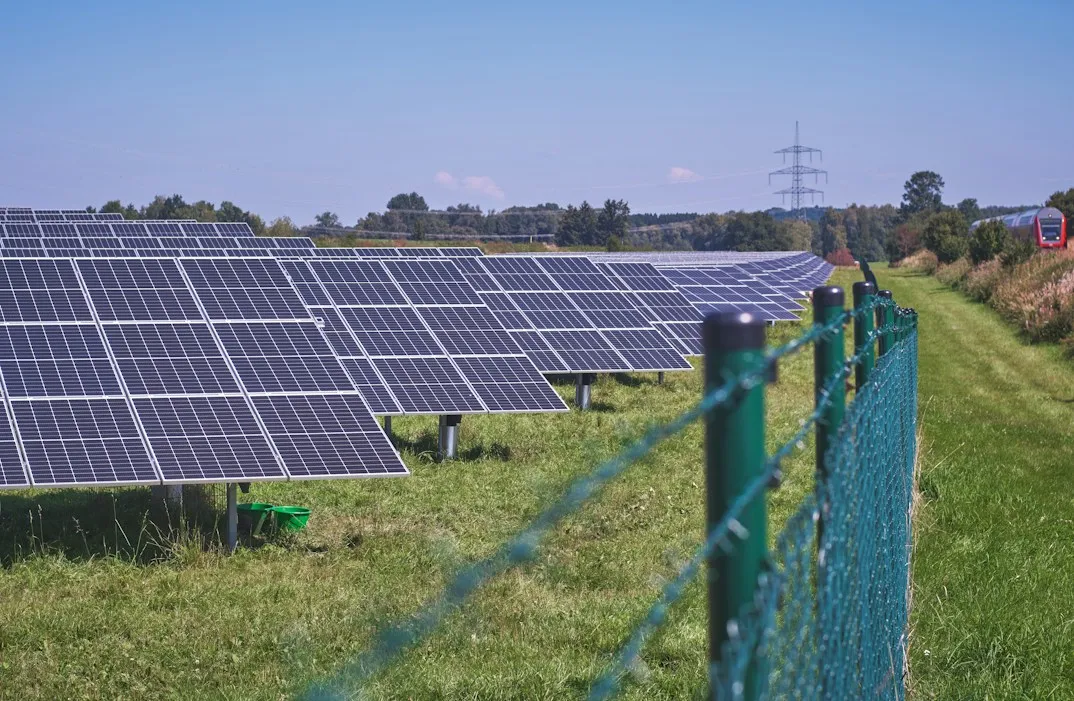 Rows of solar panels in a grassy field behind a green fence